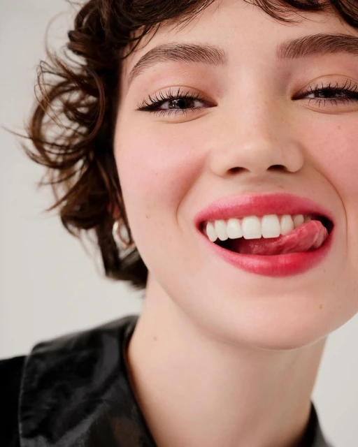 Close-up portrait of a smiling adult woman with short curly hair, red lipstick, visible tongue, wearing a black jacket against a white background.