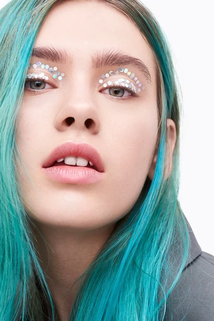 Close-up studio shot of a woman with teal hair and decorative glitter around her eyes, looking directly at the camera on a white background.