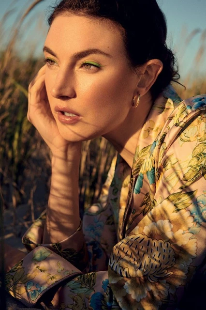 Portrait of an adult woman with green eyeshadow wearing a floral shirt in a grassy field during golden hour, looking slightly away.