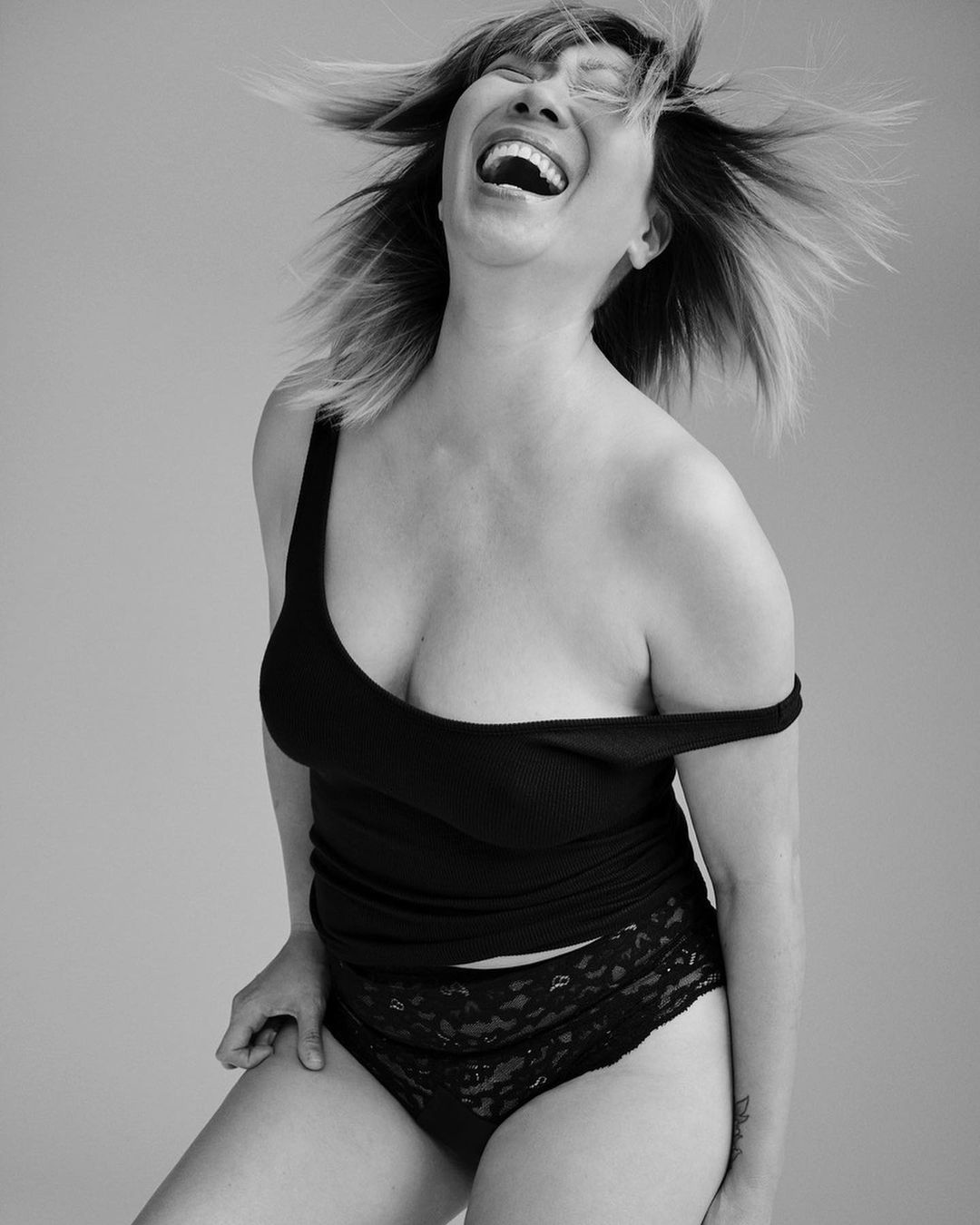 Black and white shot of a smiling adult female wearing a black tank top and lace underwear. The woman has short hair and is indoors.