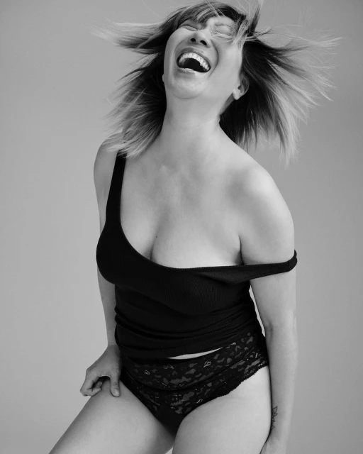 Black and white shot of a smiling adult female wearing a black tank top and lace underwear. The woman has short hair and is indoors.