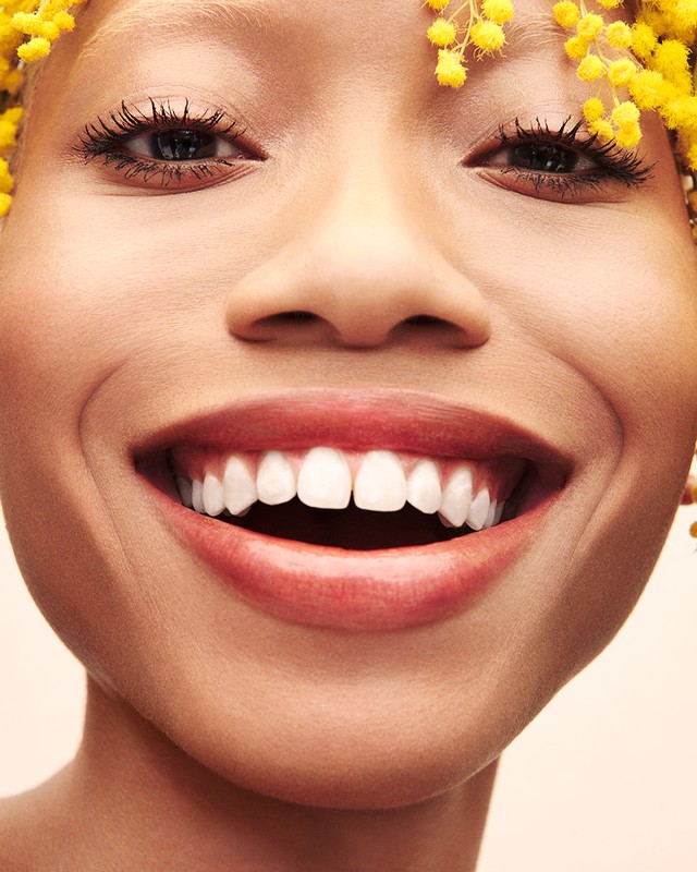 Close-up portrait of a smiling woman with white teeth and yellow flowers in her hair against a neutral background, full-frame shot.