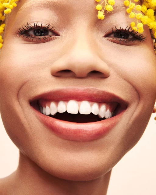 Close-up portrait of a smiling woman with white teeth and yellow flowers in her hair against a neutral background, full-frame shot.