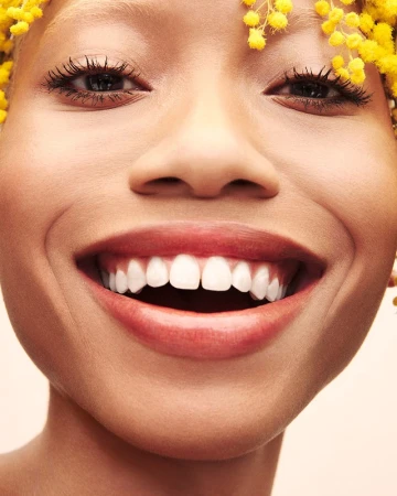 Close-up portrait of a smiling woman with white teeth and yellow flowers in her hair against a neutral background, full-frame shot.