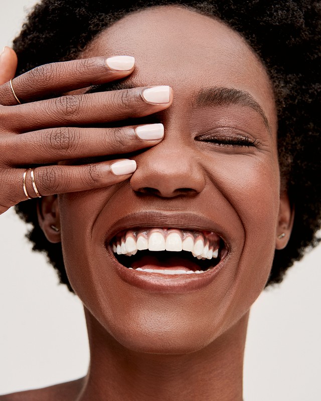 Close-up portrait of a smiling Black woman covering her eyes with her hand showing light pink manicured fingernails.