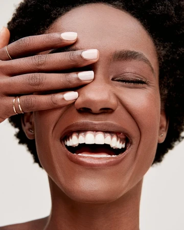 Close-up portrait of a smiling Black woman covering her eyes with her hand showing light pink manicured fingernails.