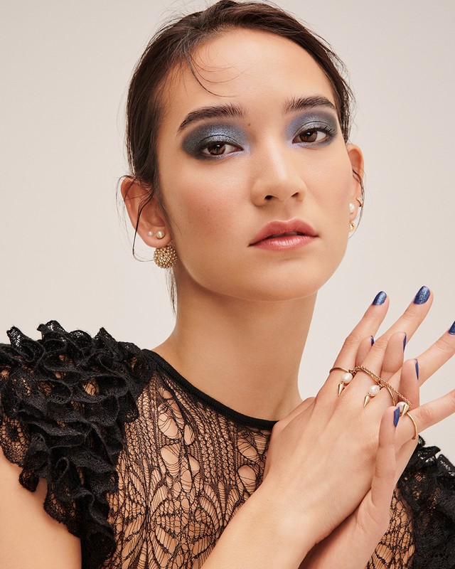 Close-up of a woman with dark hair and smokey eye makeup wearing a black lace top and statement rings in a neutral setting.