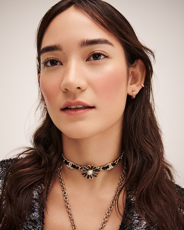 Close-up of a woman with dark hair wearing a black-and-gold flower choker and a gold chain necklace against a light background.
