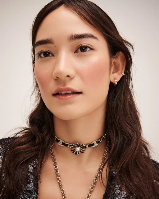 Close-up of a woman with dark hair wearing a black-and-gold flower choker and a gold chain necklace against a light background.