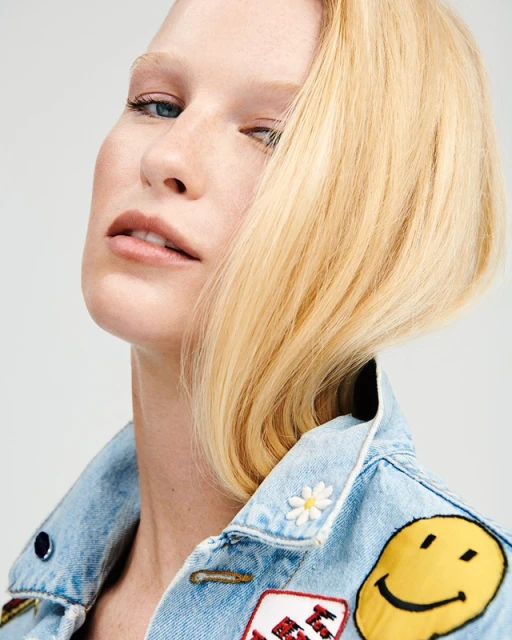 Woman with blonde hair poses in a denim jacket decorated with buttons and patches against a neutral background in a portrait shot.