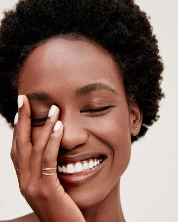 Close-up portrait of a smiling Black woman with short afro hair holding her hand near her face with white manicured nails.