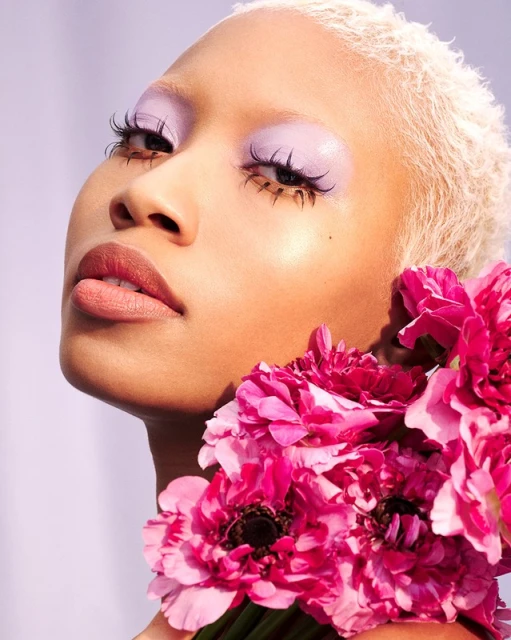 Close-up of a model with bleached blonde hair and lavender eyeshadow next to bright pink flowers, against a pale lavender backdrop.
