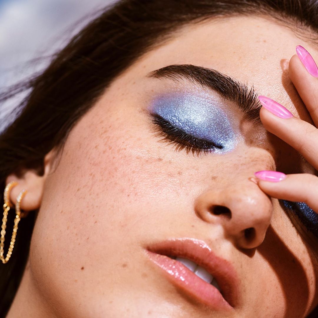 Close-up portrait of a fair-skinned adult female model with bright blue eye shadow and glossy lips, posing with hand near face.