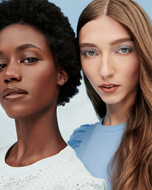 Two women, one African American and one Caucasian, stand closely together, looking at the camera in a beauty or fashion shot.