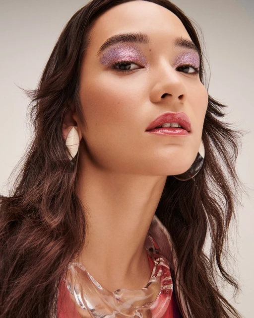 Close-up of a young woman with pink glitter eyeshadow and clear, sculptural necklace, studio shot. Beauty and fashion advertising.