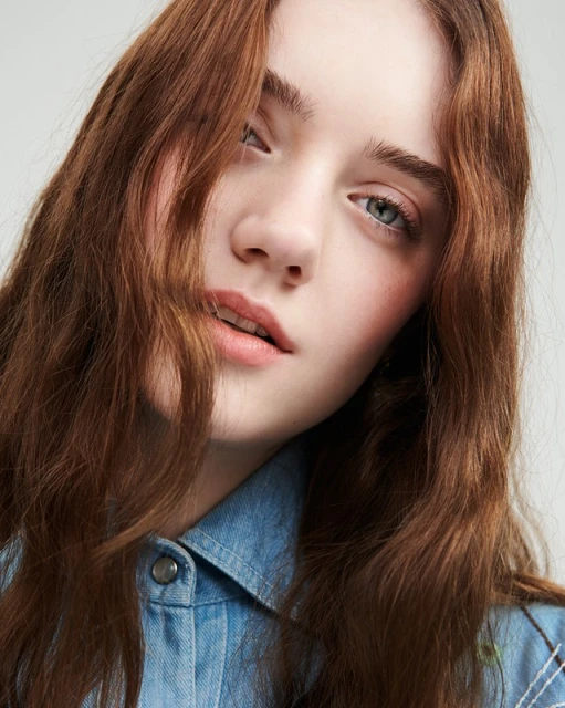 Close-up portrait of a young woman with auburn wavy hair wearing a denim shirt against a neutral studio background.
