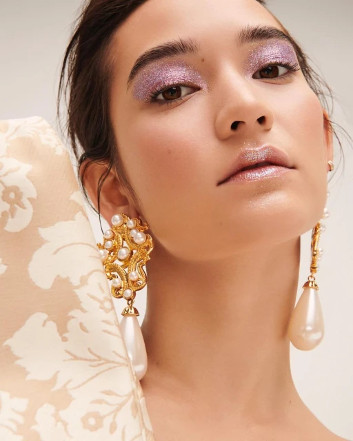 Close-up of a woman with glitter eyeshadow and statement pearl earrings, posing in a studio with floral pattern textile.