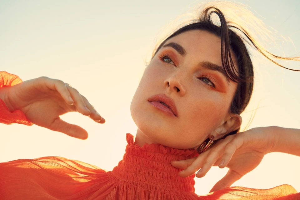Portrait of a woman with warm makeup and orange dress, looking upward in a sunlit outdoor setting, creating a soft and dreamy atmosphere.