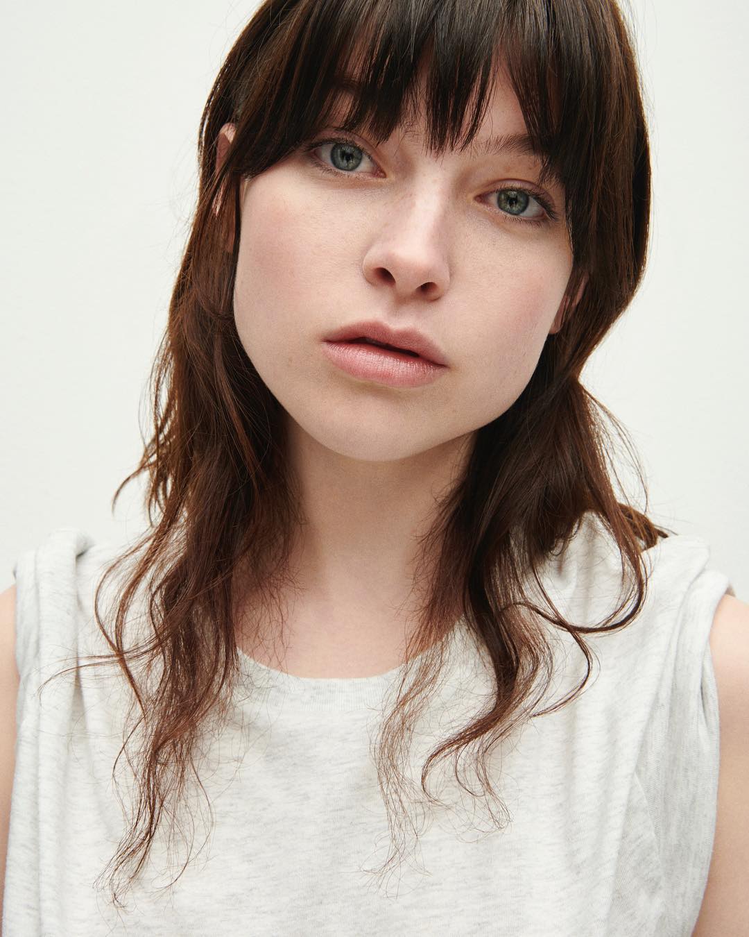 Close-up portrait of an adult woman with wavy brown hair and a grey shirt against a clean, neutral background with soft, diffused lighting.