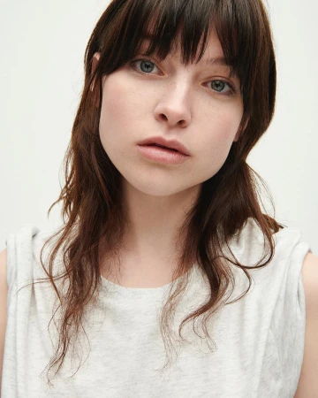 Close-up portrait of a young woman with dark hair and bangs wearing a light grey sleeveless top against a plain white background.