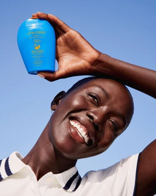 Happy adult woman holds Shiseido sunscreen bottle up against blue sky, smiling in a beauty shot emphasizing skincare and sun protection.