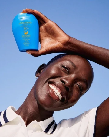 Happy adult woman holds Shiseido sunscreen bottle up against blue sky, smiling in a beauty shot emphasizing skincare and sun protection.