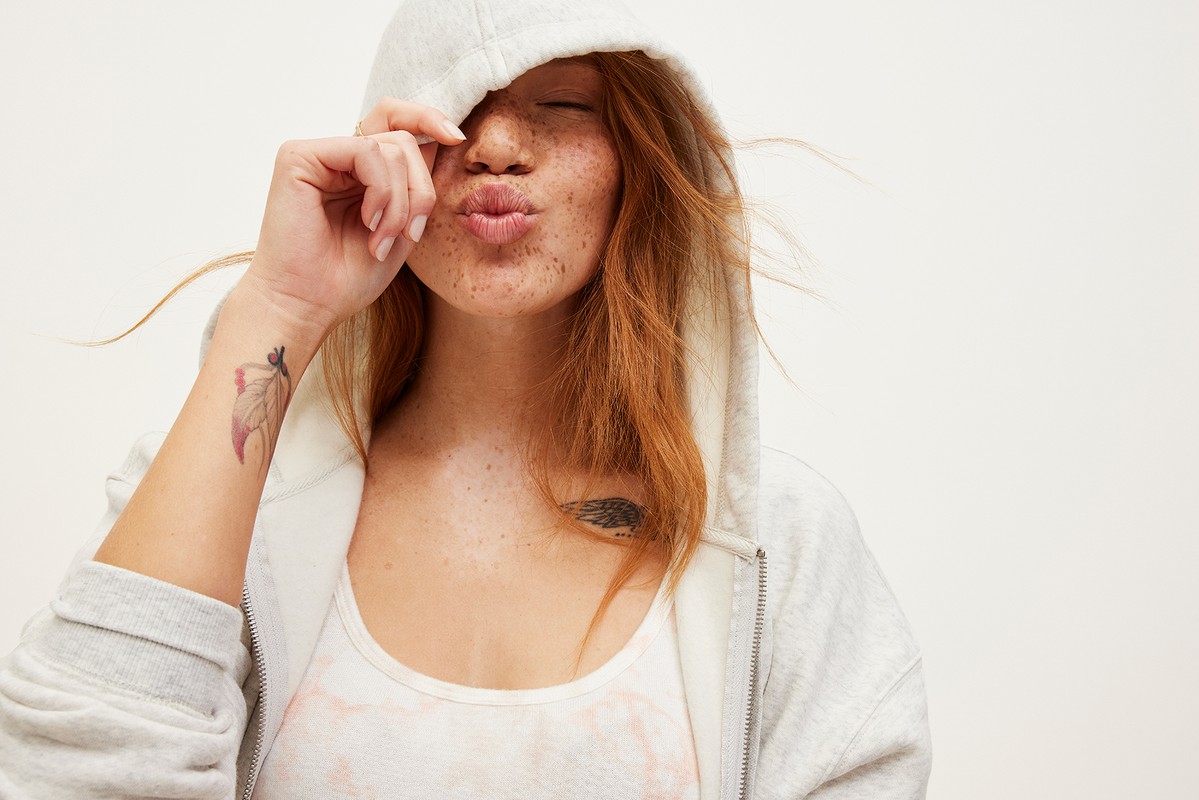A young fair-skinned woman with red hair and freckles poses in a light grey zip hoodie, puckering her lips for the camera.