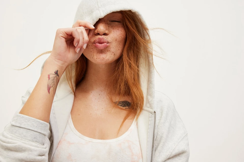 A young fair-skinned woman with red hair and freckles poses in a light grey zip hoodie, puckering her lips for the camera.