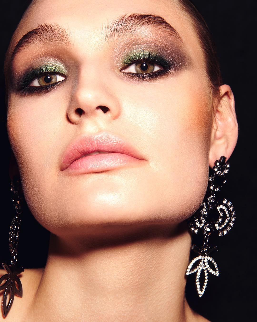 Close-up of a woman with olive green eyeshadow, pink lipstick, and long black chandelier earrings against a dark background.