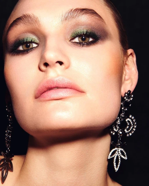 Close-up of a woman with olive green eyeshadow, pink lipstick, and long black chandelier earrings against a dark background.