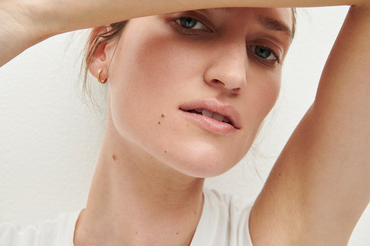 Close-up studio shot of a young adult female with fair skin, gold hoop earrings, and a white shirt posing against a white backdrop.