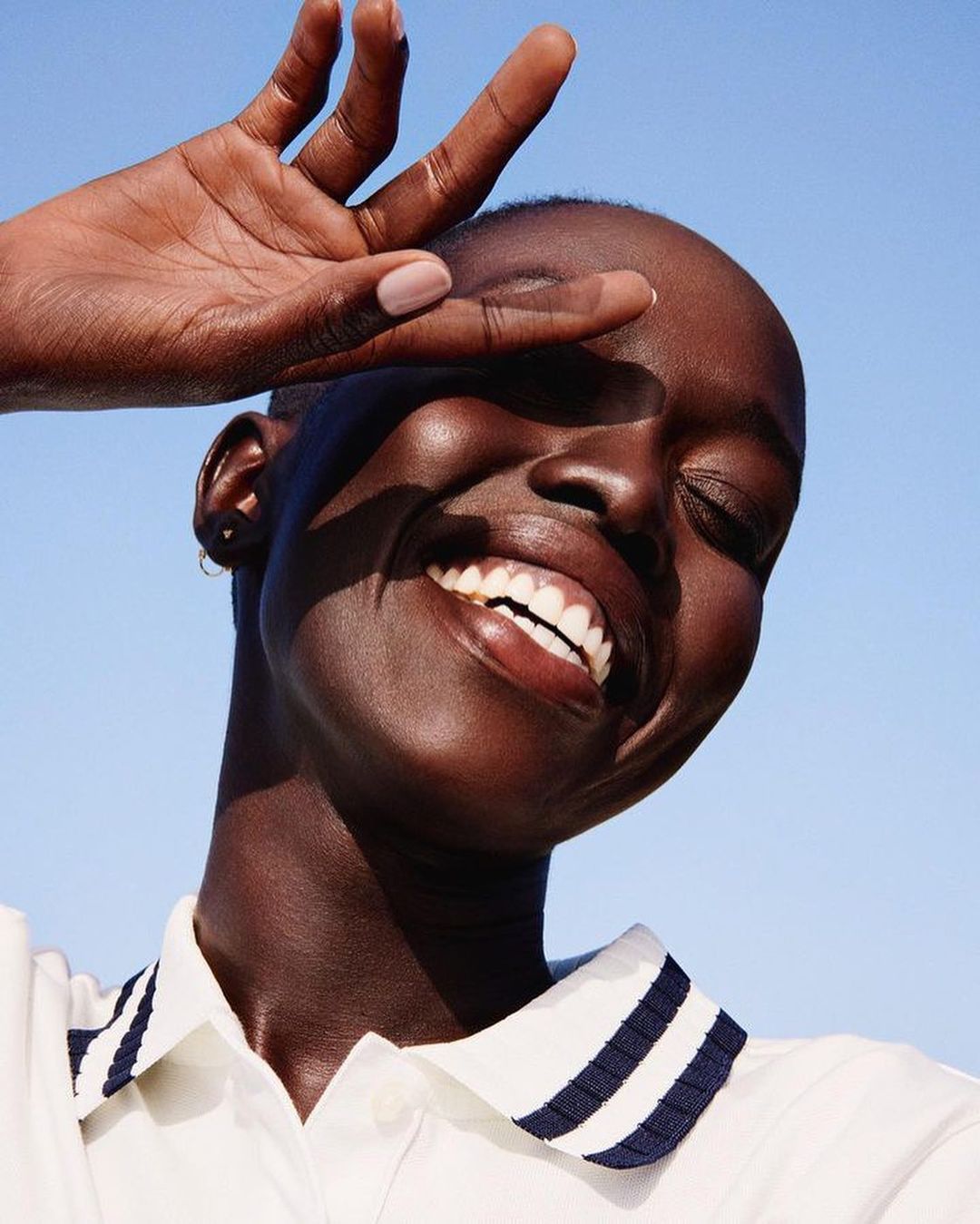 Close-up portrait of a smiling Black woman with hand shielding eyes against bright blue sky, wearing a white shirt with navy collar.