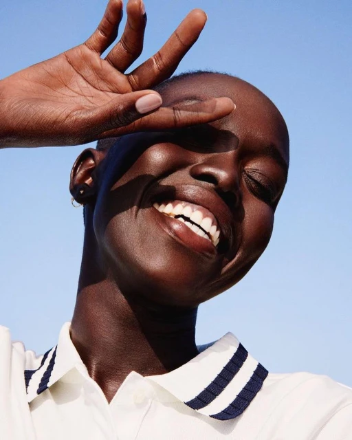Close-up portrait of a smiling Black woman with hand shielding eyes against bright blue sky, wearing a white shirt with navy collar.