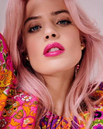 Close-up portrait of a woman with pink hair wearing bright pink lipstick and a colourful patterned top.