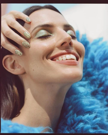 Close-up portrait of a woman smiling with closed eyes, wearing pale green eyeshadow, patterned nails, and a textured blue garment.
