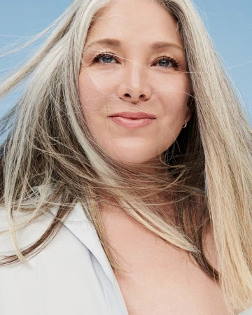 Close-up portrait of an adult woman with long gray hair against a light blue background, showcasing natural beauty and healthy aging.