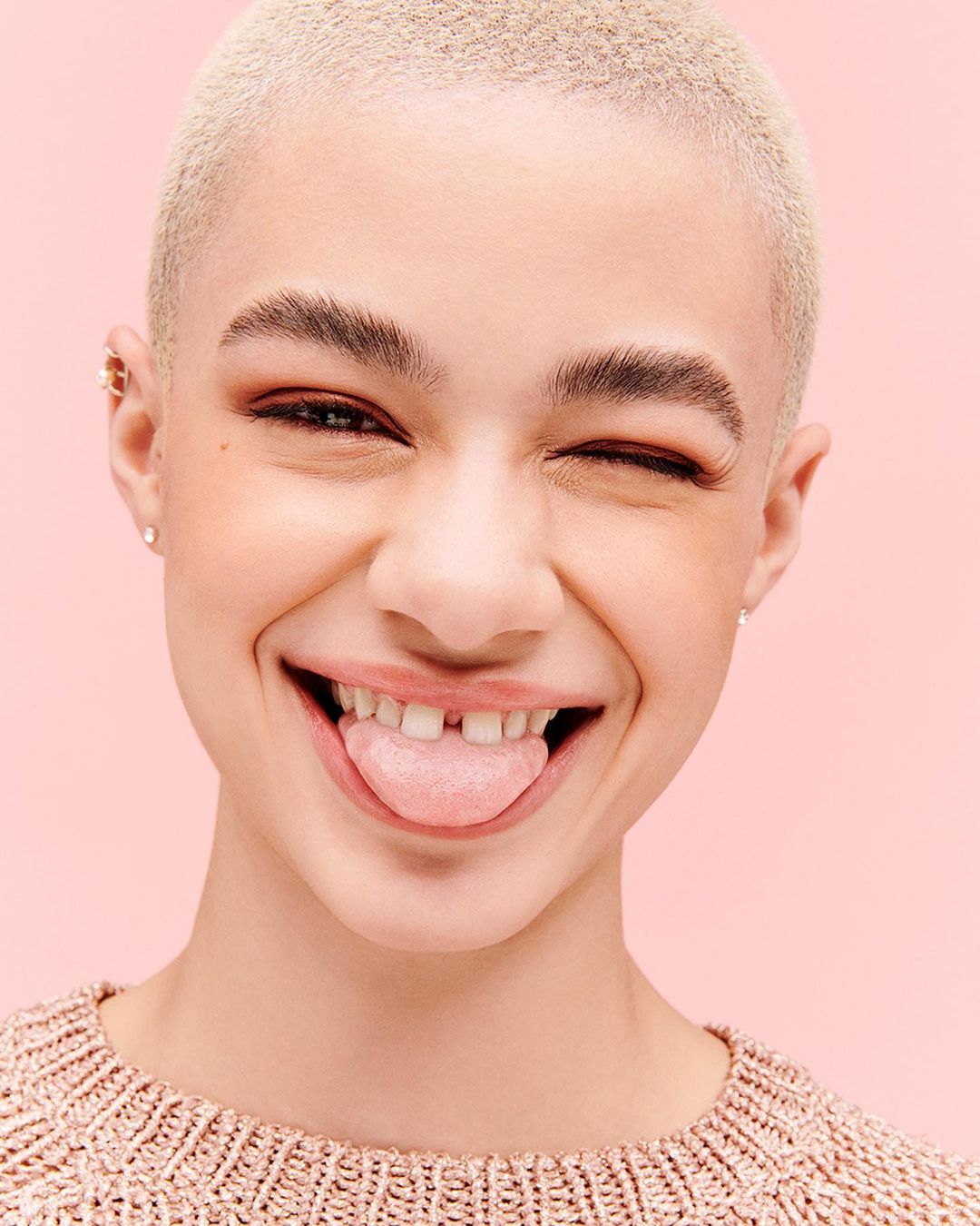 Close-up of a young adult female with a shaved head, fair complexion, and a warm, bright smile sticking her tongue out on a pink background.