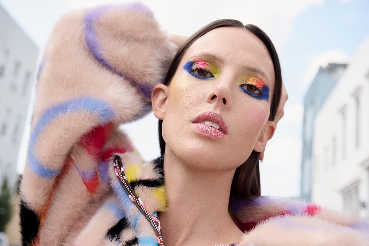 Close-up of a woman with colorful eyeshadow and a fur coat, posing in an urban environment with buildings in the background.