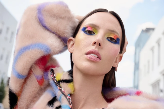 Close-up of a woman with colorful eyeshadow and a fur coat, posing in an urban environment with buildings in the background.
