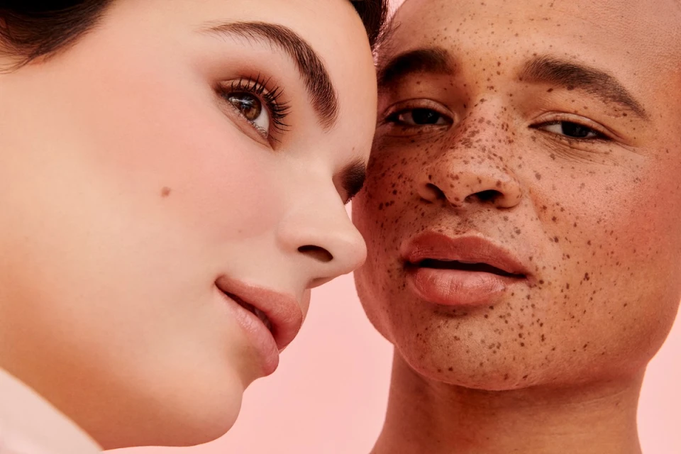 Close-up shot of faces of two adults, one with fair skin and one with freckled skin, in a beauty or skincare product advertisement on pink backdrop.