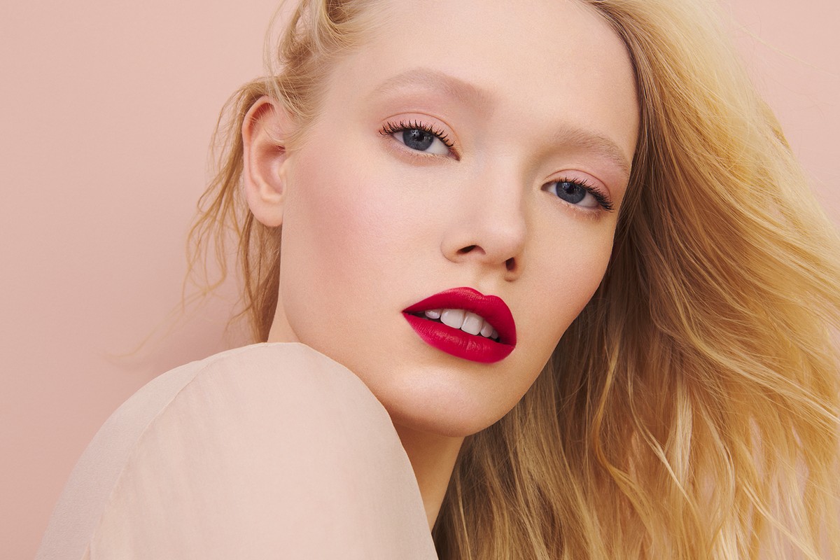 Close-up portrait of a blonde woman with red lipstick posing against a pink backdrop in a beauty and fashion context.