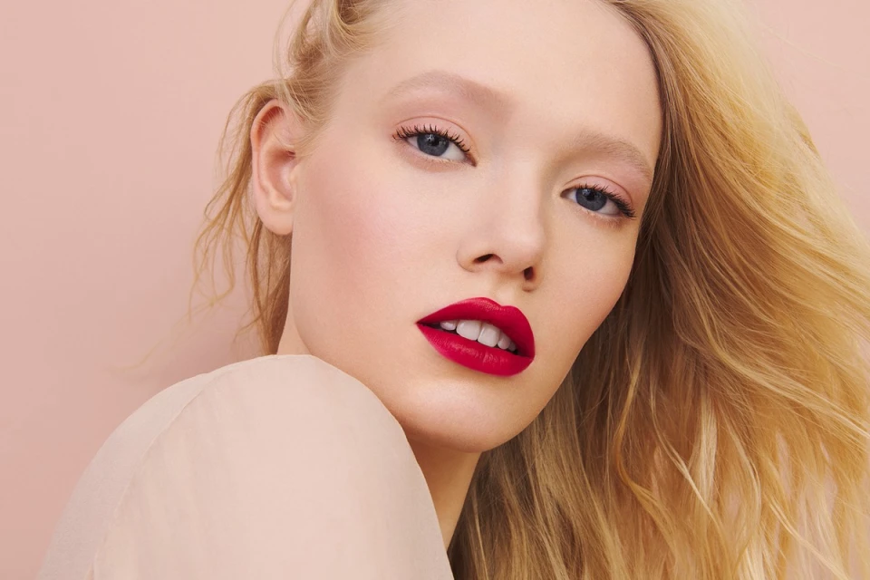 Close-up portrait of a blonde woman with red lipstick posing against a pink backdrop in a beauty and fashion context.