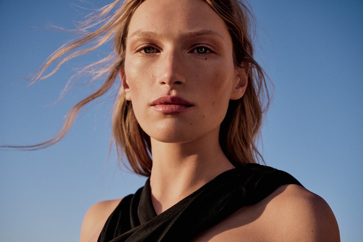 Close-up portrait of a woman with long blonde hair blowing in the wind wearing a black top against a clear blue sky
