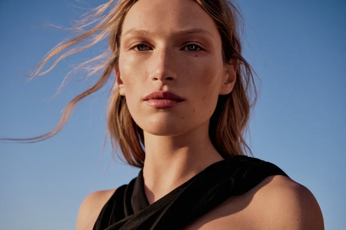 Close-up portrait of a woman with long blonde hair blowing in the wind wearing a black top against a clear blue sky