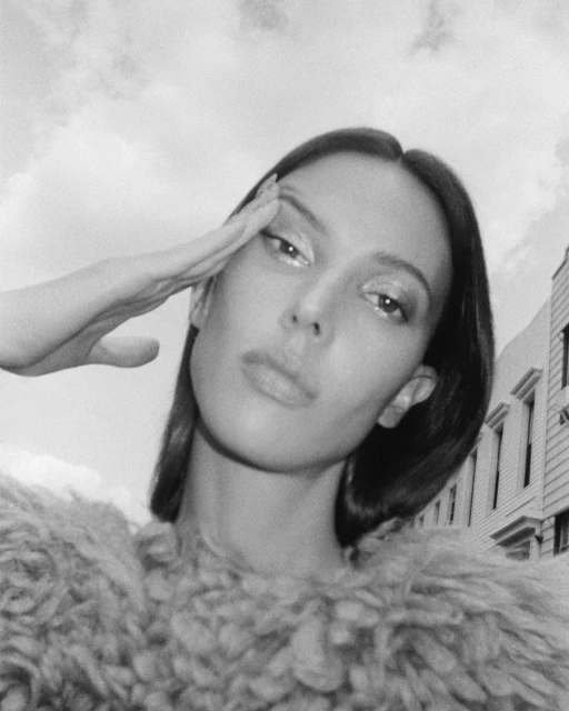 Black and white portrait of a woman in a textured top with hand raised to her forehead, with a city street and cloudy sky behind.