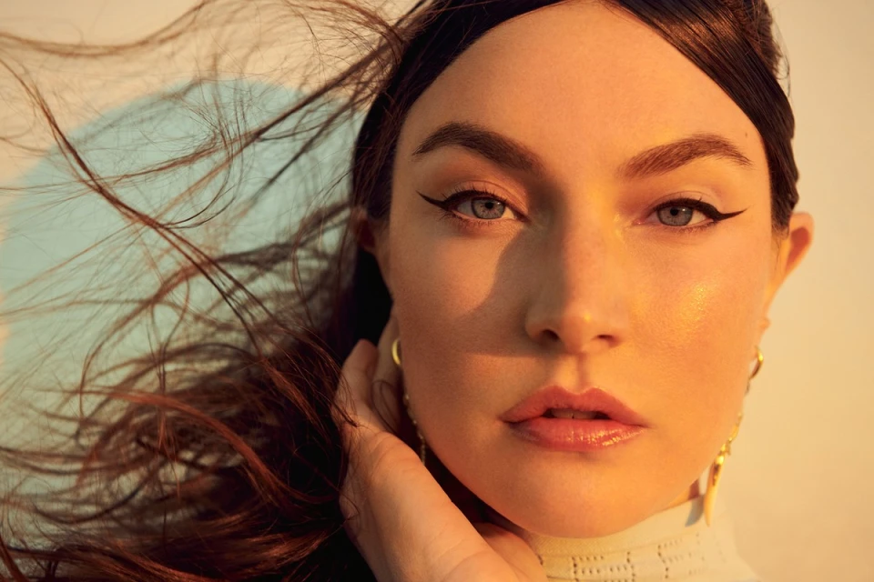 Close-up portrait of a woman with dark hair blowing in the wind, featuring makeup, gold earrings, and a textured collar in warm lighting.