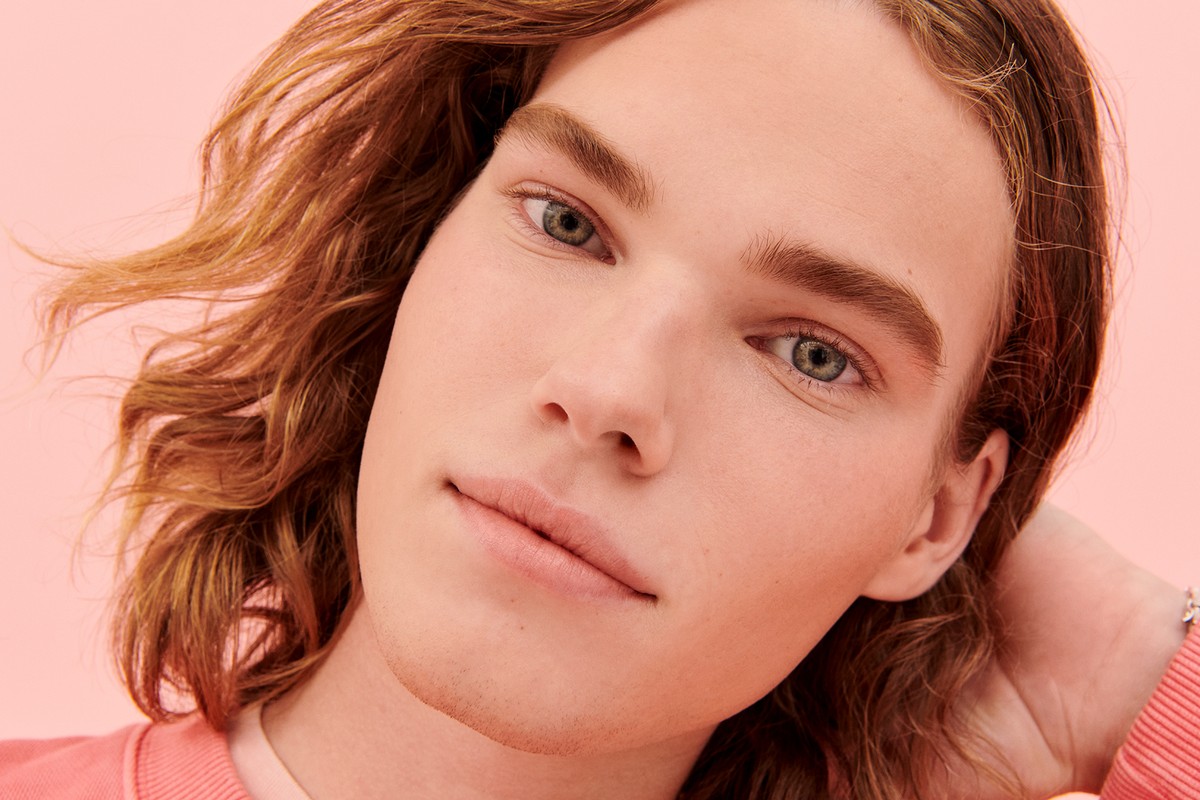 Close-up portrait of a fair-skinned young adult with long blonde hair, styled in a modern beauty aesthetic, against a light peach backdrop.