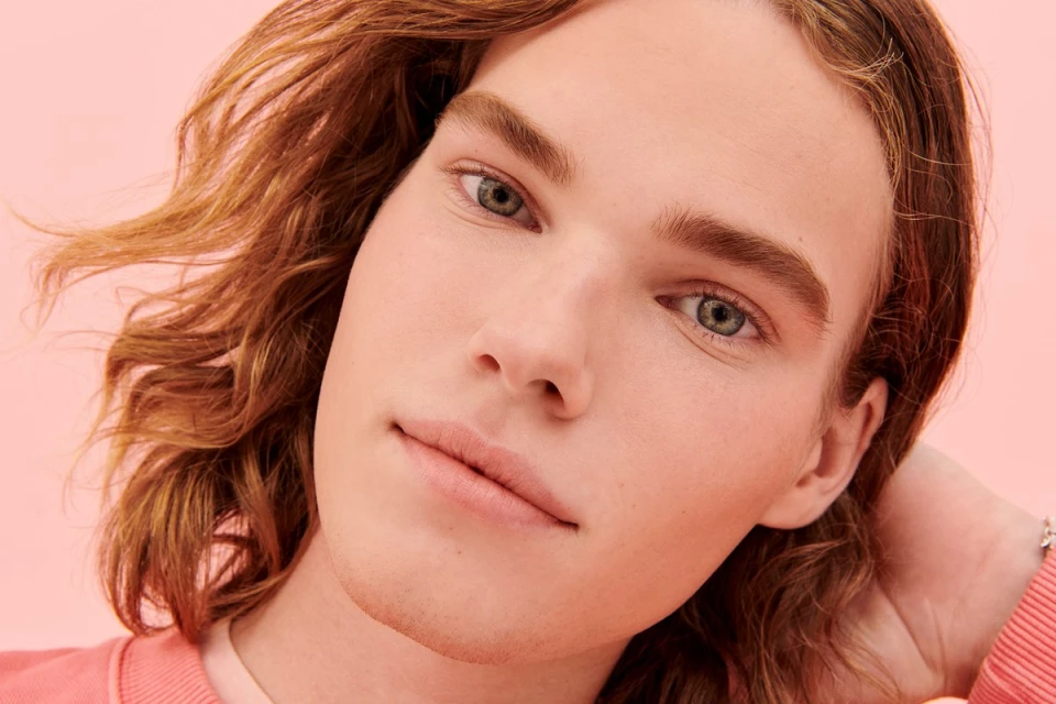 Close-up portrait of a fair-skinned young adult with long blonde hair, styled in a modern beauty aesthetic, against a light peach backdrop.