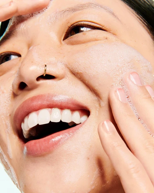 Close-up of a smiling Asian woman washing her face with soapy foam, her fingers gently massaging her skin, showcasing skincare and beauty.
