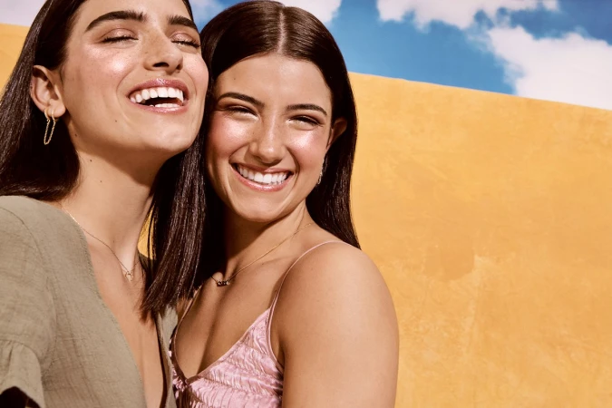 Two smiling young women with dark hair posing close together against a warm yellow and blue sky background